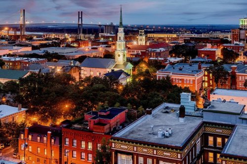 Aerial view of a city at dusk, featuring historic buildings, a church steeple, and a bridge in the background.