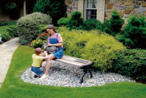 A child kneels beside a seated woman on a bench, holding a rabbit in a lush garden with colorful flowers.