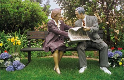 A bronze sculpture of a couple sitting on a bench, reading a newspaper together amidst colorful flowers.
