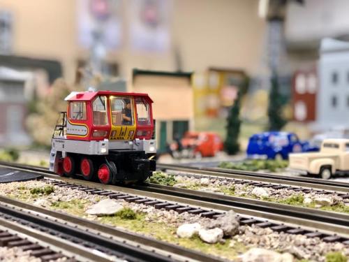 A model train engine on a miniature railway, surrounded by small buildings and vehicles in a detailed landscape.