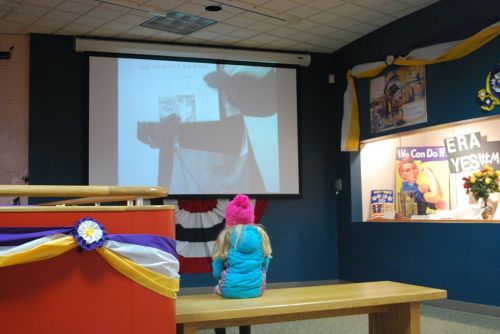 A child in a pink hat watches a presentation on a screen in a decorated room.