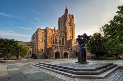 Historic stone building with a tall tower, surrounded by trees and a modern sculpture in the foreground.