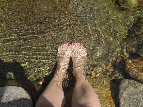 A pair of feet with red nail polish submerged in clear, rippling water over smooth stones.