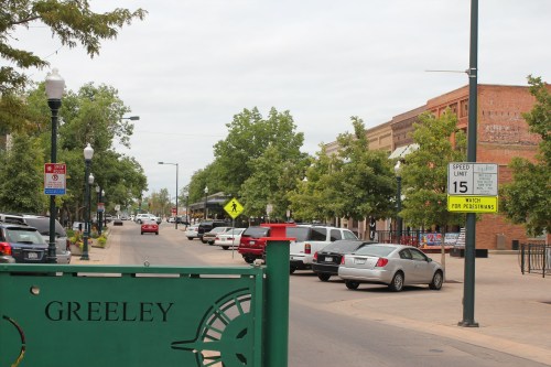 A street view of Greeley, featuring trees, parked cars, and signage for pedestrians and speed limits.