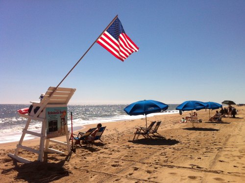 A sunny beach scene with a lifeguard stand, American flag, and beachgoers under umbrellas on the sand.