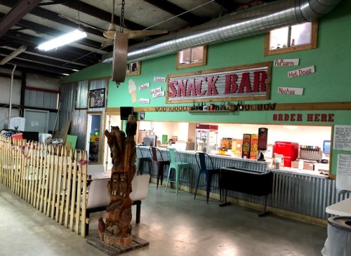 A rustic snack bar with colorful decor, seating, and a menu featuring popcorn, hot dogs, and nachos.