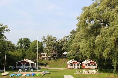 A scenic view of red cabins on a grassy area, surrounded by trees and kayaks, under a clear blue sky.