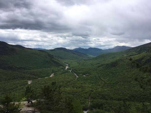 A panoramic view of lush green valleys and mountains under a cloudy sky. A winding river flows through the landscape.