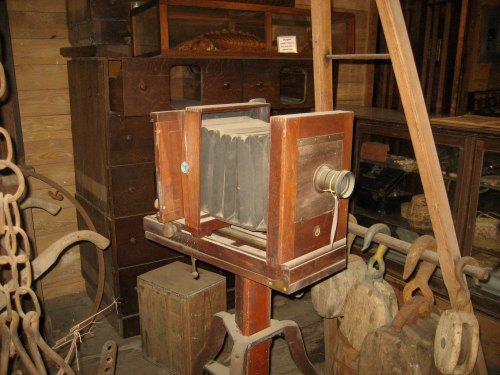 An antique wooden camera on a stand, surrounded by vintage furniture and tools in a rustic setting.