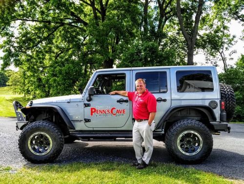 A man in a red shirt stands next to a silver Jeep with "Penn's Cave" branding in a green outdoor setting.