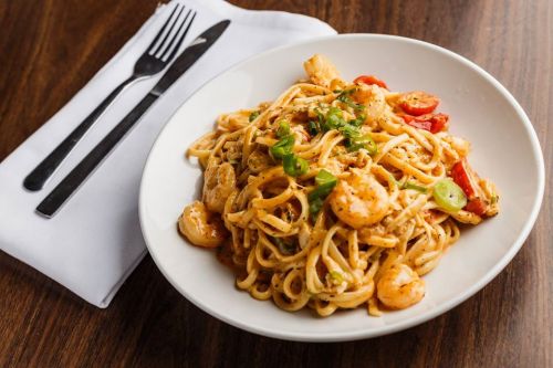 A plate of shrimp pasta with green onions and tomatoes, served on a wooden table with a fork and knife beside it.