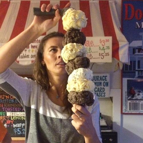 A woman balances a tall stack of ice cream scoops on a cone, looking up in surprise at her creation.