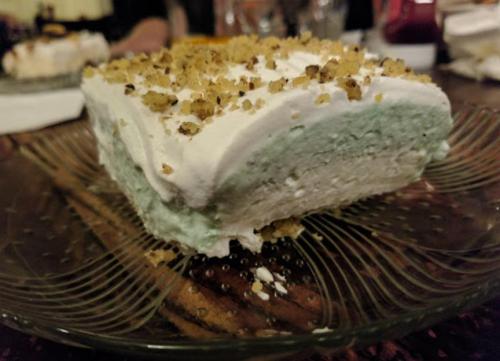 A slice of layered dessert with a creamy green filling, topped with whipped cream and crushed nuts on a decorative plate.