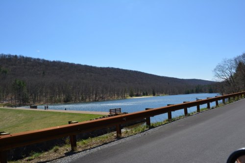 A serene lake surrounded by trees and hills, with a clear blue sky and a road running alongside it.