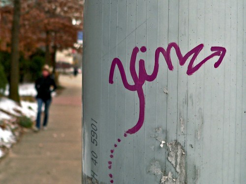 A close-up of purple graffiti on a pole, with a blurred figure walking in the background on a snowy sidewalk.