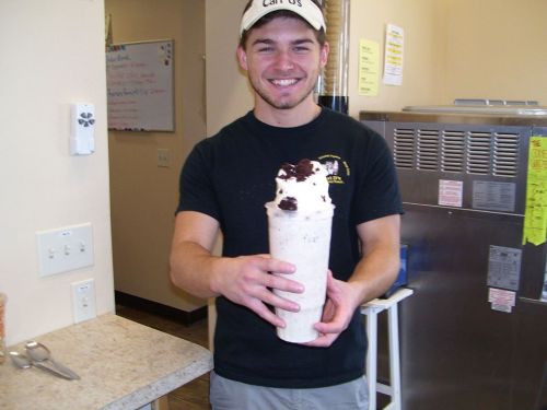 A smiling young man holds a large milkshake topped with whipped cream and cookies, standing in an ice cream shop.