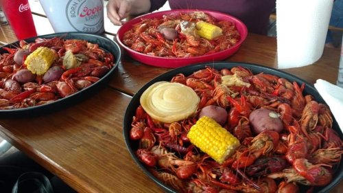 Two large platters of crawfish with corn, potatoes, and a bun, served on a wooden table.