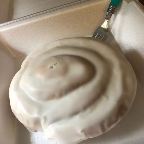 A large cinnamon roll with white icing, placed in a takeout container with a fork beside it.