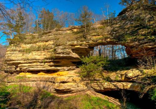 A natural rock formation with an arch, surrounded by trees and blue sky, showcasing rugged terrain and greenery.