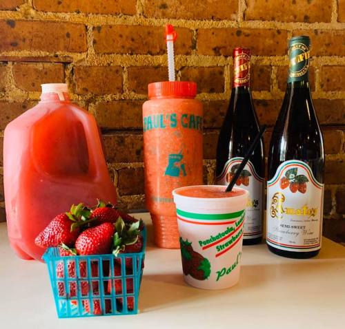 A colorful display of strawberry drinks, a basket of fresh strawberries, and bottles of wine against a brick background.