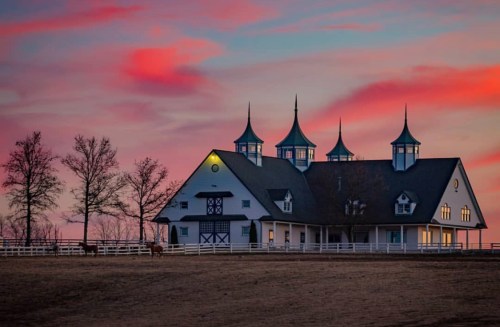 A picturesque barn with spires at sunset, surrounded by trees and a fence, with horses grazing nearby.