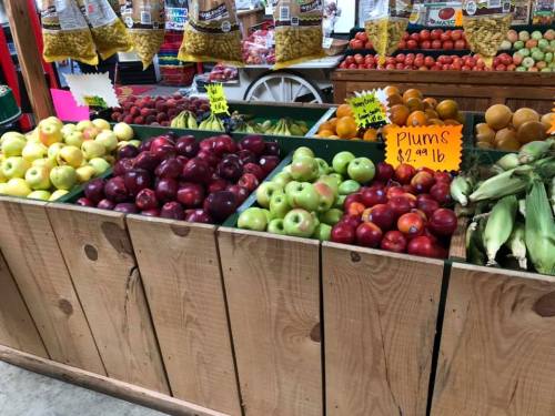 A wooden display filled with various apples, plums, and corn at a market, with colorful signs and bags of snacks in the background.