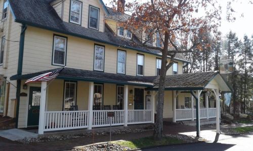 A charming yellow inn with a porch, rocking chairs, and an American flag, surrounded by trees on a sunny day.