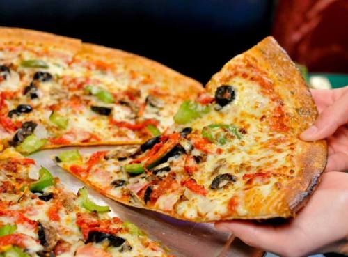 A hand reaching for a slice of pizza topped with olives, peppers, and meats, on a pizza tray.