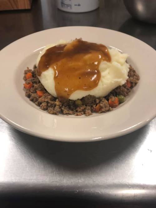A plate of mashed potatoes topped with gravy, served over a bed of ground meat mixed with carrots and peas.