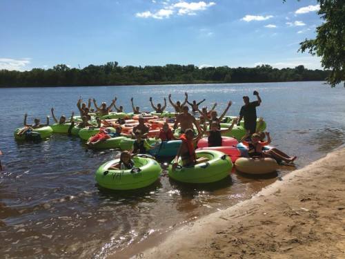 A group of people on colorful inner tubes in a river, enjoying a sunny day and celebrating with raised arms.