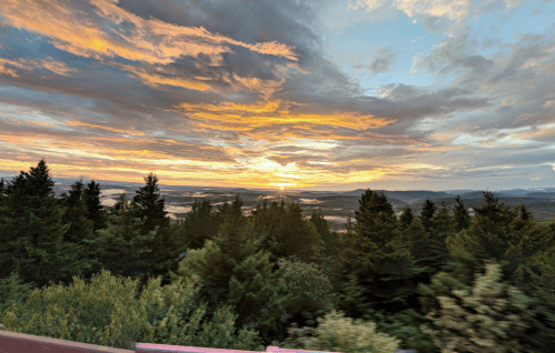 A vibrant sunset over a valley, with lush green trees in the foreground and colorful clouds in the sky.