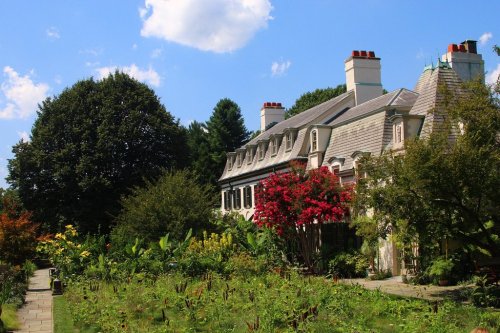 A charming house with a sloped roof, surrounded by vibrant gardens and trees under a blue sky with fluffy clouds.