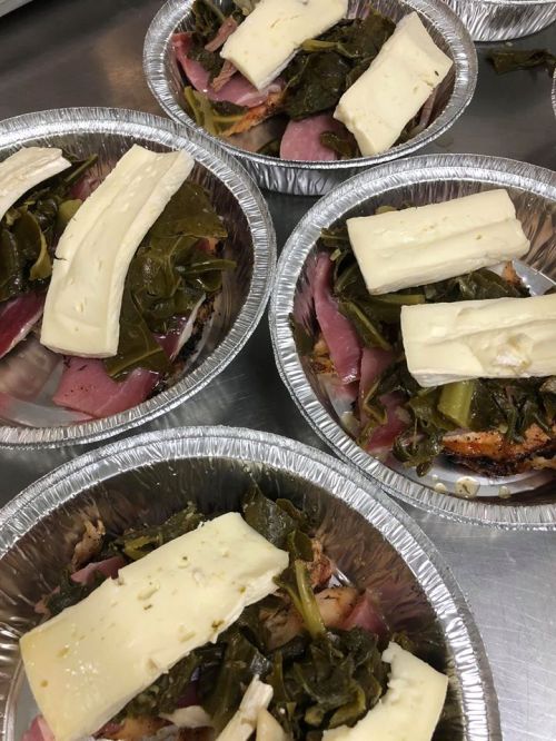 Four aluminum trays filled with greens, ham, and topped with slices of cheese.