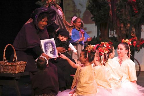 A performer in a purple costume reads to children in costumes, surrounded by festive decorations on stage.