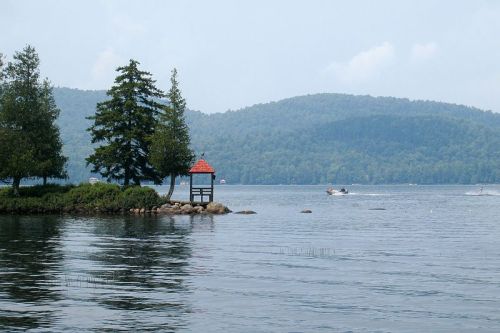 A serene lake scene with trees, a small gazebo, and a boat on the water, surrounded by green hills under a cloudy sky.