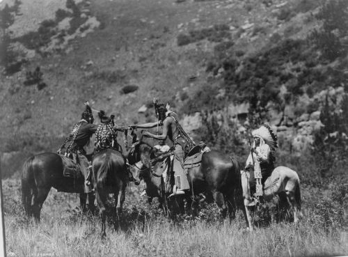 A historical black-and-white photo of Indigenous people on horseback in a natural landscape, wearing traditional attire.