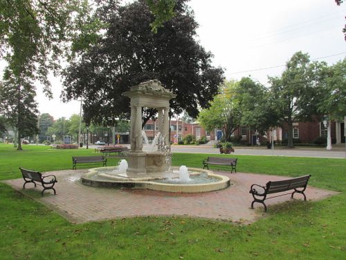 A decorative fountain surrounded by benches in a green park, with trees and a street visible in the background.