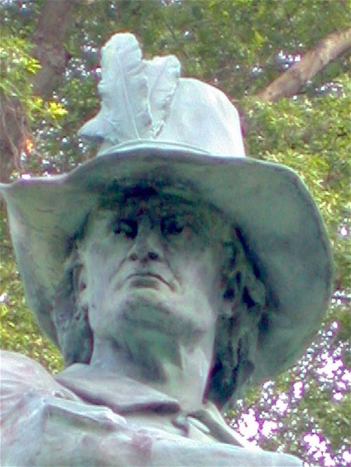 Close-up of a bronze statue of a man wearing a wide-brimmed hat with a feather, surrounded by greenery.