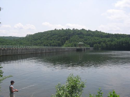 A person fishing by a calm lake with a dam and lush green hills in the background under a partly cloudy sky.