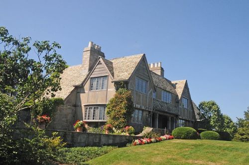 A large, elegant house with a stone exterior, multiple chimneys, and a well-maintained garden under a clear blue sky.