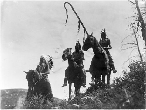Two Native American riders on horseback, one holding a lasso, against a mountainous backdrop. Black and white photo.