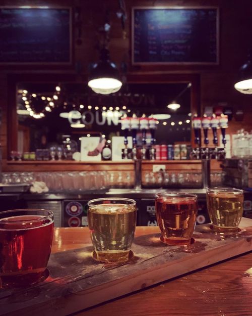 A wooden flight board with four colorful shot glasses in a cozy bar setting, illuminated by warm lights.