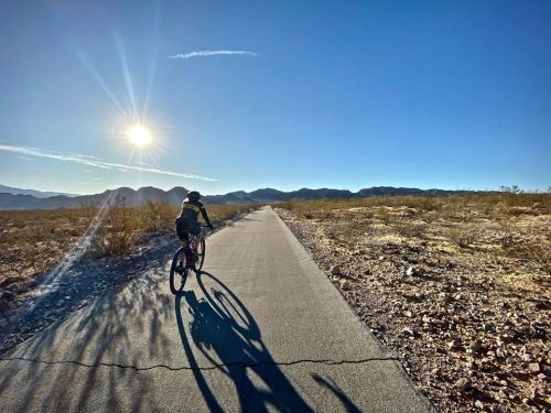 A cyclist rides on a paved path through a desert landscape, with mountains and a bright sun in the background.
