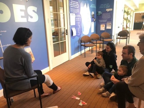 A woman reads to a small group of children and adults sitting on the floor in a colorful, educational space.
