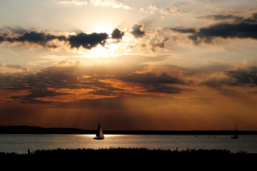 A sailboat glides across a calm lake at sunset, with dramatic clouds and rays of sunlight illuminating the sky.
