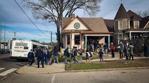 A group of people gathers outside a building, with a bus parked nearby and trees in the background.