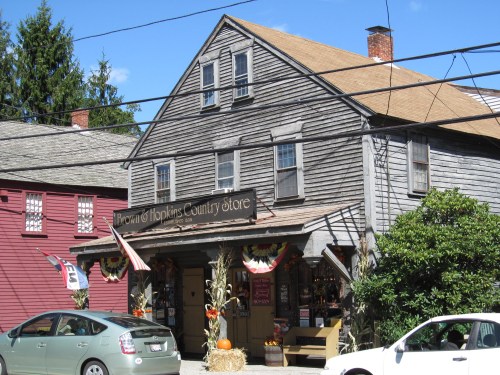 A rustic country store with a wooden facade, decorated for fall, surrounded by trees and a parked car.