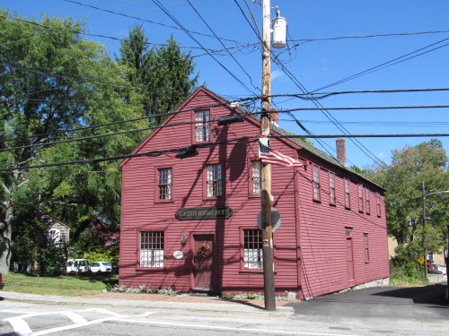 A red historic building at a street corner, surrounded by trees and power lines, under a clear blue sky.