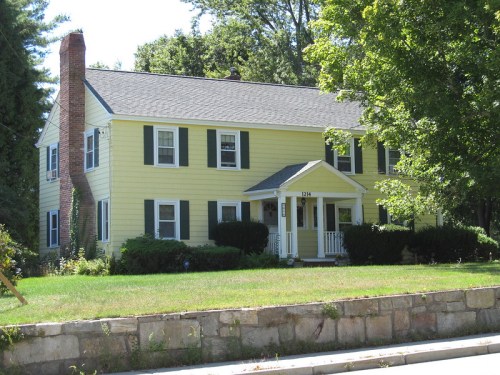 A two-story yellow house with green shutters, a front porch, and a well-maintained lawn surrounded by trees.