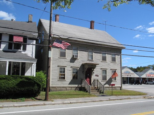 A two-story wooden house with an American flag, surrounded by greenery and shops in the background under a clear blue sky.